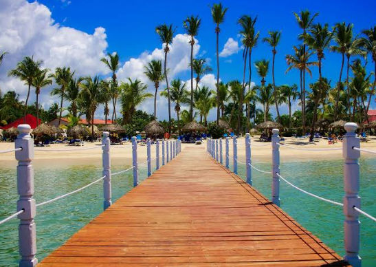 Wooden boardwalk over clear blue water leading to a tropical beach with palm trees and resort amenities.