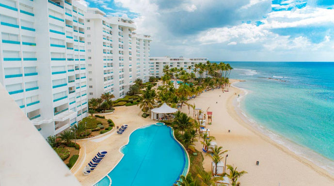 Vista aérea de resort frente al mar con edificios blancos, piscina azul y palmeras