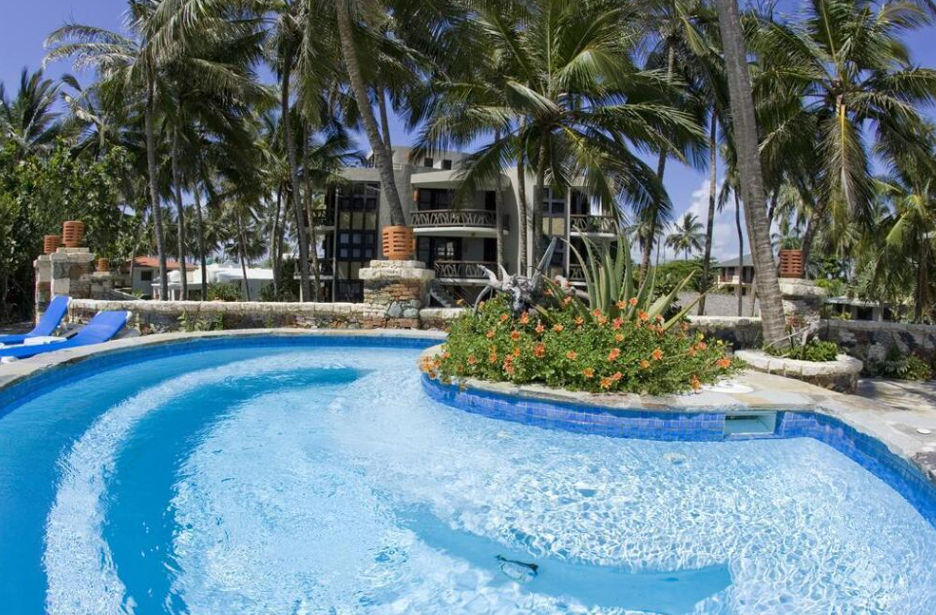 A sunny daytime photograph of a tropical resort swimming pool, with six blue lounge chairs lined up. Behind the pool are tall coconut palm trees, dense lush foliage, and a multi-story building with balconies under a clear blue sky.