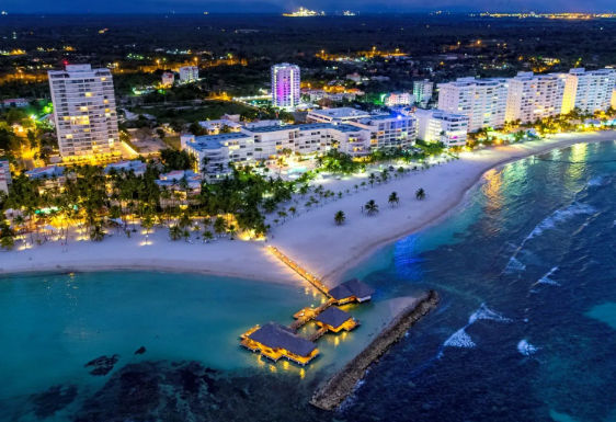 Aerial night view of a luxury beach resort in the Dominican Republic, with illuminated hotels and a wooden pier with overwater bungalows.