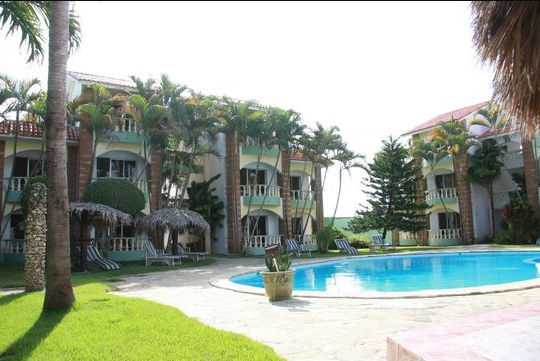 A nighttime photograph of a multi-story resort complex surrounding an illuminated swimming pool. The light yellow buildings feature numerous balconies and brick accents. Tall palm trees, smaller landscaping plants, and several lounge chairs surround the pool area under a deep blue evening sky.