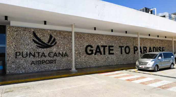 Exterior of Punta Cana Airport with a stone wall reading Gate to Paradise and a silver minivan parked outside