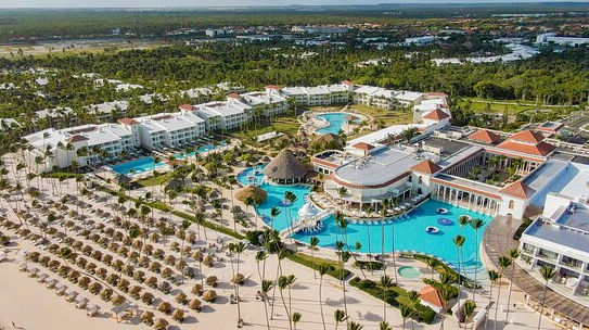 Aerial view of a luxurious all-inclusive resort in Punta Cana, Dominican Republic, featuring swimming pools, a white sand beach with palapas, and lush palm trees.