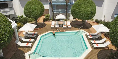 An aerial view of a landscaped resort complex, featuring multi-story buildings with distinctive blue roofs surrounding a winding, lagoon-style swimming pool. The pool area is bordered by a patterned red and white tiled deck and includes a central thatch-roofed palapa and a white gazebo. The property is filled with lush greenery, stone-lined paths, green lawns, and several unique spherical topiary trees, under a blue sky with wispy clouds.
