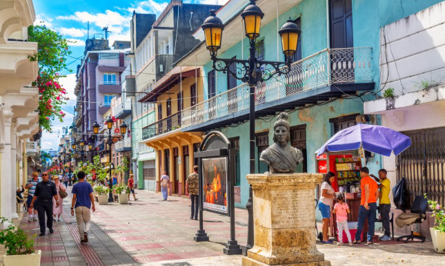 Vibrant colonial street scene with colorful buildings, pedestrians, a bronze bust statue, and classic street lamps