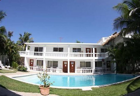 Daytime view of a modern two-story white and grey hotel building surrounding a rectangular blue swimming pool. The hotel features balconies with classic white balustrade railings and sliding glass doors. In the foreground, two modern chaise lounge chairs rest on a wooden deck next to the pool. A rooftop terrace with a covered area is on the right, and green palm trees frame the top background against a clear blue sky.