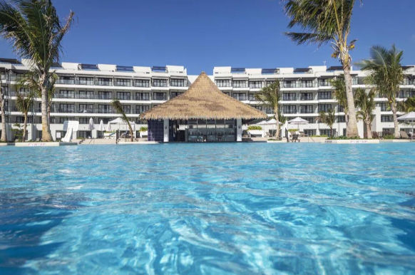 Large resort swimming pool with a thatched-roof swim-up bar and modern white hotel buildings in the background