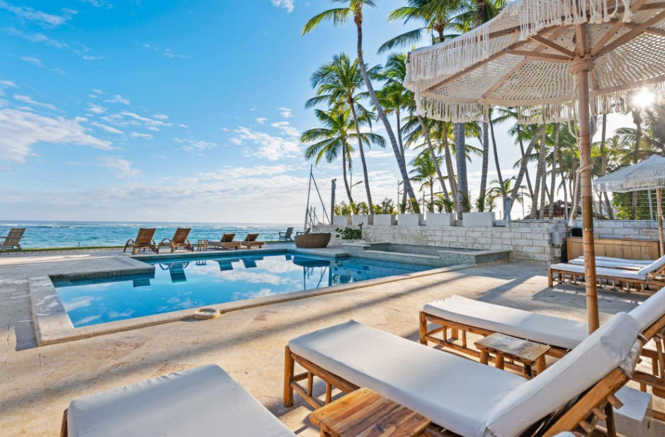 A beachfront swimming pool area with white lounge chairs, a fringed umbrella, tall palm trees, and the ocean under a sunny blue sky.