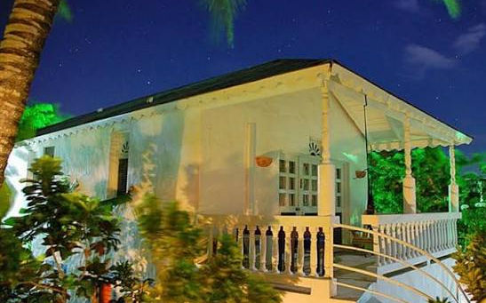 Casa tropical blanca con porche de noche, rodeada de plantas verdes iluminadas bajo un cielo estrellado.