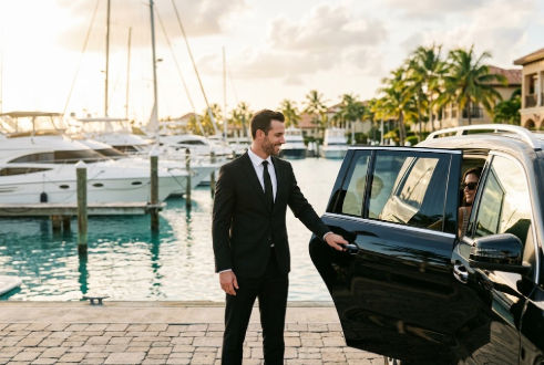 Chauffeur opens the door of a luxury SUV at a marina with yachts and palm trees.