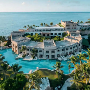 Aerial view of a castle-style beachfront resort with a large blue swimming pool and palm trees