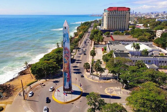 Vista aérea de un malecón costero con una avenida principal, edificios de la ciudad y un gran obelisco decorado con murales junto al mar