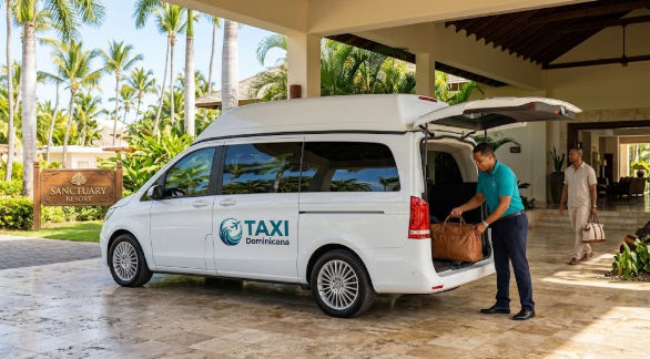 A male attendant in a blue polo shirt loading luggage into the trunk of a white TAXI Dominicana passenger van at the entrance of Sanctuary Resort with palm trees.