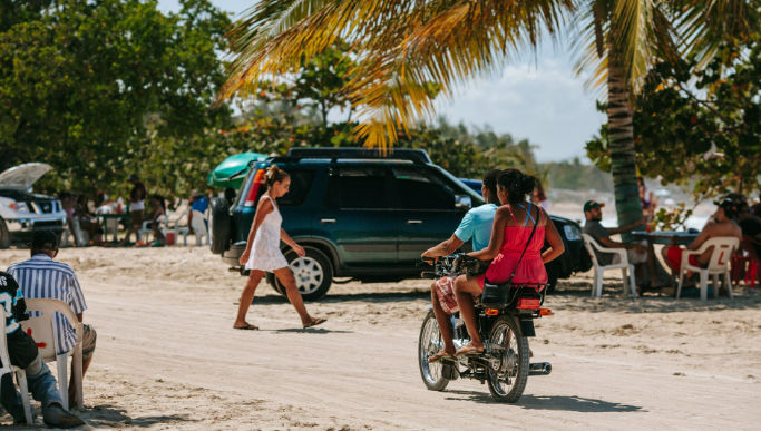 A man and woman riding a motorbike on a sandy path at Macao Beach with an SUV and palm trees.