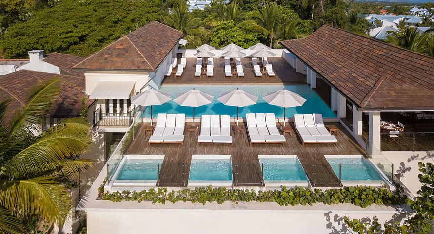 Aerial photograph of a luxurious resort swimming pool area. A woman floats in the large turquoise main pool on the right. To the left, a wooden deck features several white lounge chairs and a series of open white patio umbrellas. Next to the deck are three smaller, separate square plunge pools.