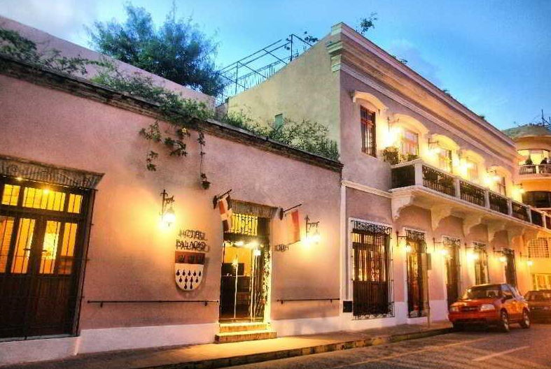 Vista exterior nocturna iluminada del histórico Hotel Palacio en una calle adoquinada, mostrando secciones de fachada rosa y beige, balcones de hierro forjado adornados con luces cálidas, el letrero del "HOTEL PALACIO" y un escudo de armas