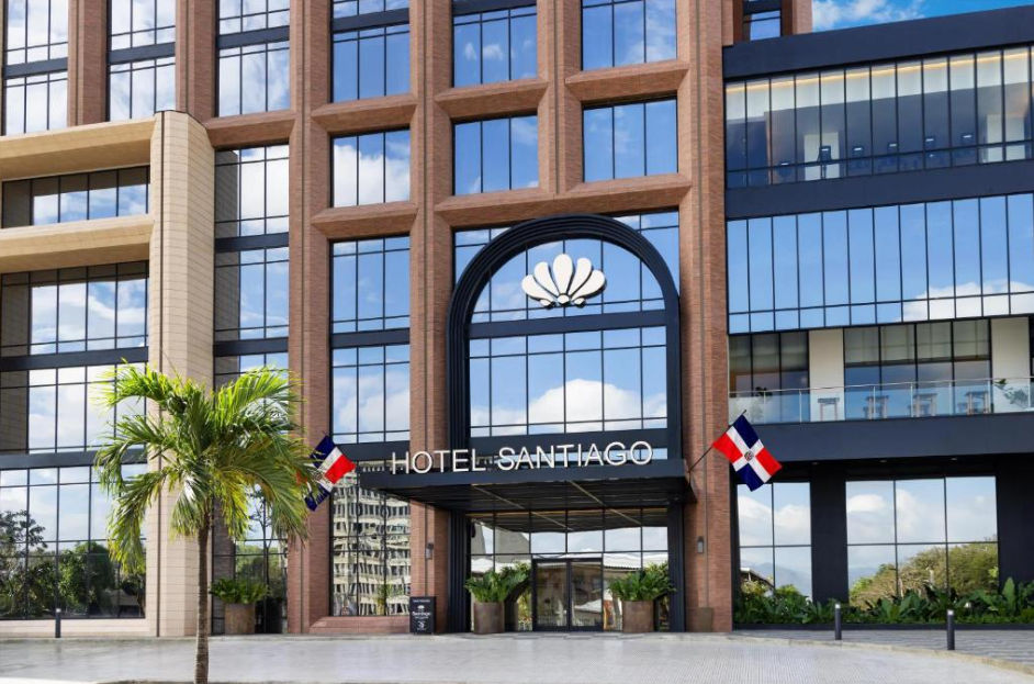 La entrada exterior del moderno Hotel Santiago de varios pisos, con fachada de ladrillo rojo y vidrio azul reflectante. El marco arqueado negro de la entrada tiene un logotipo de concha, y un dosel negro dice 'HOTEL SANTIAGO', flanqueado por dos banderas 