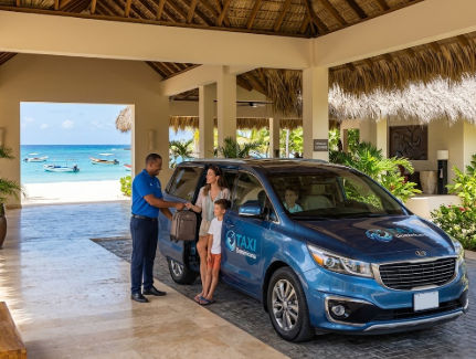 Un conductor de Taxi Dominicana ayuda a una madre e hijo a subir a una minivan azul en la entrada de un resort tropical con vista a la playa