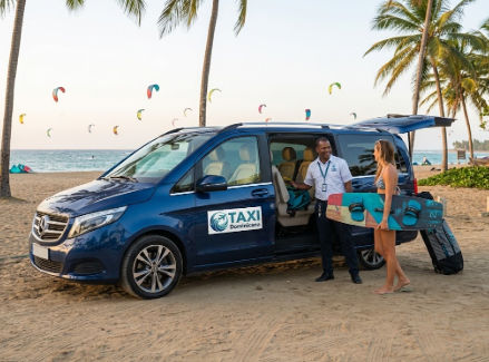 Blue Mercedes-Benz V-Class minivan taxi on Cabarete beach with a driver assisting a female tourist with a wakeboard