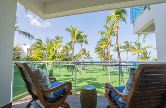 Aerial view of Seawinds at Punta Goleta beachfront resort with white buildings, a central swimming pool, expansive green lawns with palm trees, and the ocean crashing against a rocky breakwater.