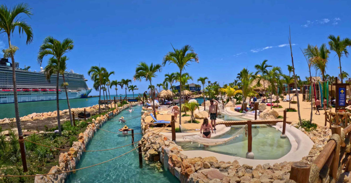Aerial view of a tropical beach club featuring a large, winding swimming pool, multiple cabanas, and large thatch-roofed palapas surrounded by palm trees, with a sandy beach facing the ocean on the right and a street with colorful buildings in the background.