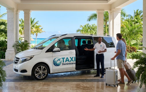 Tourists with luggage board a white Taxi Dominicana minivan at a luxury resort entrance, overlooking a tropical beach.