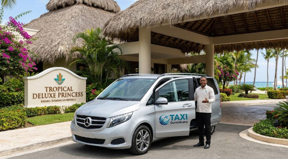 Taxi Dominicana silver passenger van parked at the entrance sign of Tropical Deluxe Princess resort in Bavaro, Dominican Republic, with a driver waiting.
