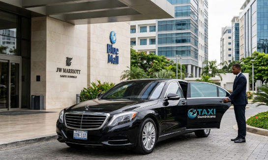 Una lujosa berlina de color negro, rotulada con 'TAXI Dominicana' y el logo de la empresa, aparcada en la entrada principal del hotel JW Marriott Santo Domingo. Un conductor uniformado mantiene la puerta trasera abierta para un cliente.