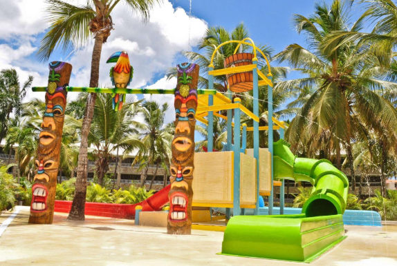 Parque acuático infantil con toboganes de colores, un cubo de agua y tótems decorativos rodeado de palmeras