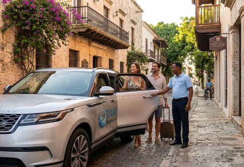Un Range Rover plateado de Taxi Dominicana con la puerta abierta, un conductor uniformado y una pareja de turistas con maleta llegando a una calle empedrada en la Zona Colonial de Santo Domingo.