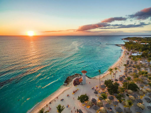 Vista aérea de la playa de Bayahibe en República Dominicana al atardecer, con un faro icónico y sombrillas de paja.