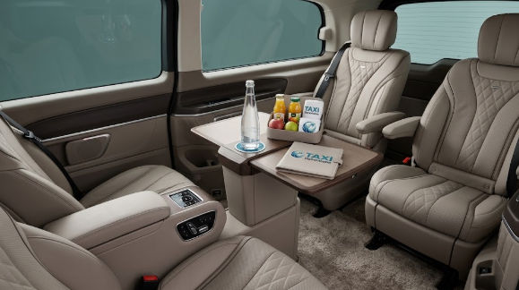 Interior de vehículo de transporte privado de lujo con asientos de cuero beige, mesa de servicio, botella de agua, frutas y folleto con la marca TAXI.