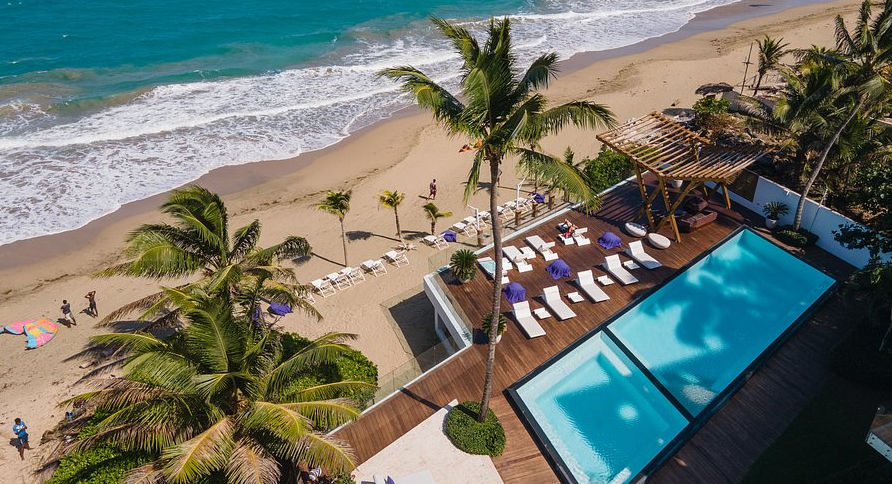 An exterior daytime photograph capturing a modern luxury resort building situated behind a large, bright blue infinity-edge swimming pool. The foreground features a wooden plank deck with several white lounge chairs and small side tables. The main building has multiple levels of balconies with glass railings and white walls, topped by a distinctive overhang roof with circular openings. Palm trees and other tropical greenery frame the building, with traditional Mediterranean-style villas and reddish-roofed buildings visible in the distance on the left under a partly cloudy sky.