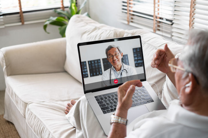 Senior man laying on couch with his laptop on lap. The laptop demonstratres a telehealth call with his doctor.