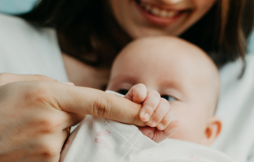 A photo of a baby being held by a female caregiver. The baby is holding the caregiver's right index finger in it's right hand. The baby is wearing a white longsleeved vest.