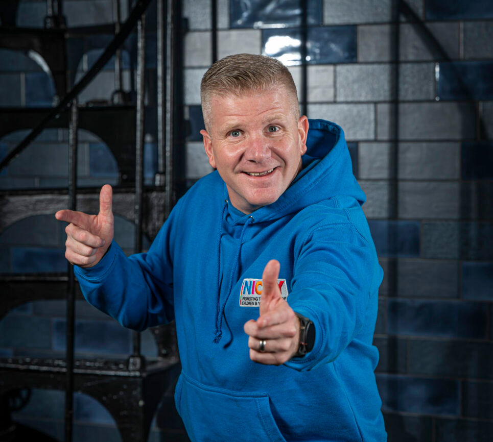 A photo of the NI Commissioner for Children and Young People, Chris Quinn smiling and playfully pointing his fingers at the camera, he is wearing a blue NICCY hoodie.