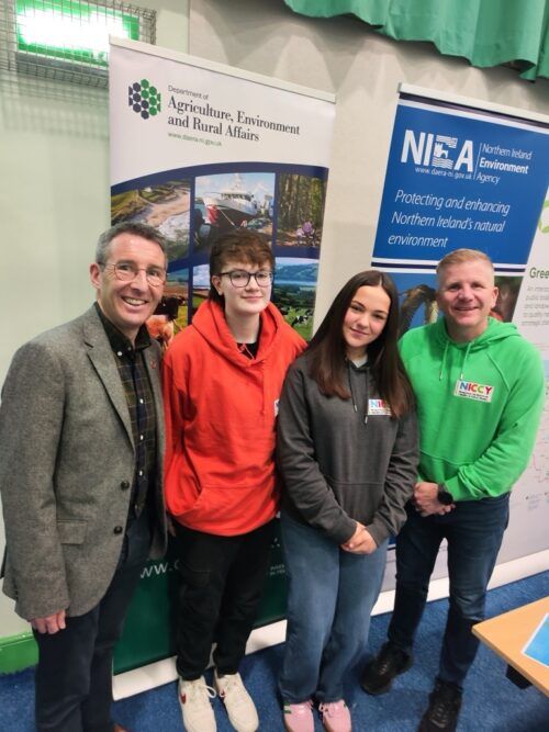 This is a photo of four people standing in a row at an event from left to right they are - DAERA Minister, Andrew Muir (MLA), NYP member Tasha, NYP member Eva, Commissioner Chris Quinn.
