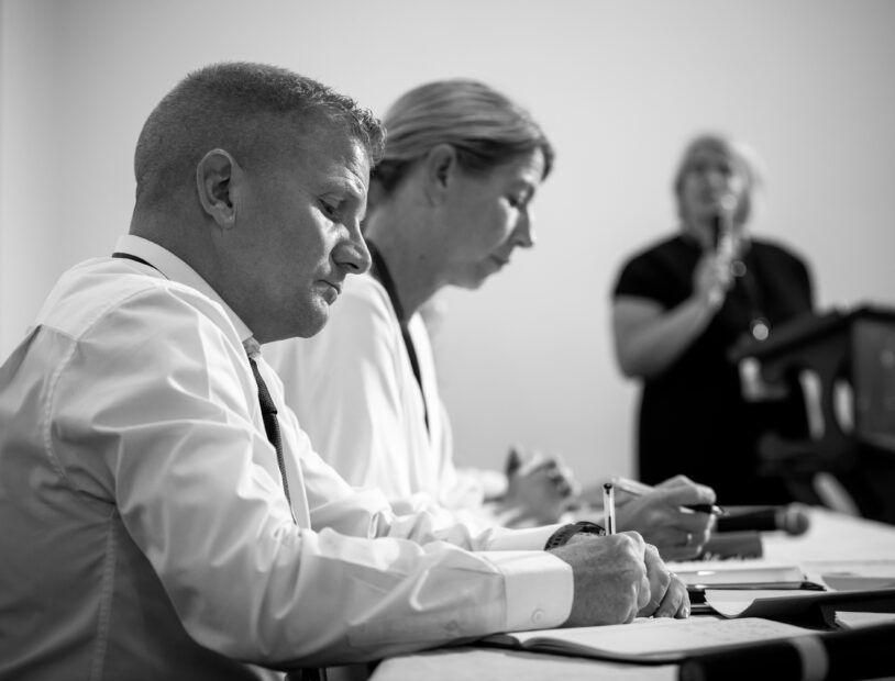 This is a black and white photo of the NI Commissioner for Children and Young People, Chris Quinn. He is sitting a desk writing a note there is a woman sitting beside him also writing a note.
