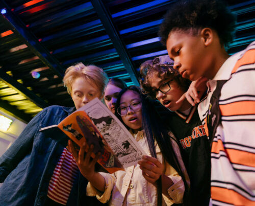 This is a photo of young children gathered round a comic book and reading it together. They are looking at the comic with amazement and awe.