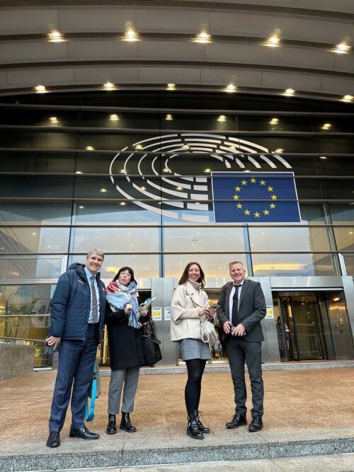 This is a photo of four ENOC delegates standing outside the glass panelled entrance of the European Parliament. It has a large European Flag (blue with a stars in a circle formation) on the front of the building. Commissioner Chris Quinn is standing at the end of the row of people on the right.