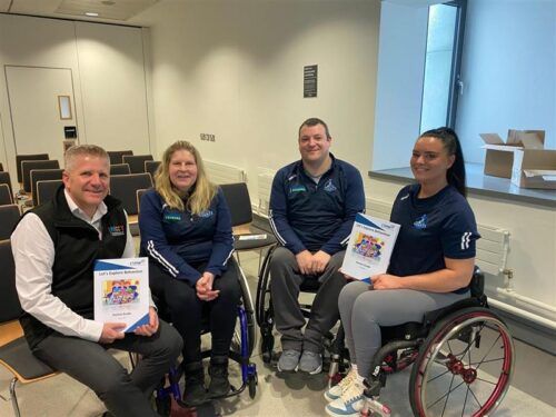 This is a photo of Commissioner Chris Quinn sitting in a row of adults that are wheelchair users. They are in a white room with a window behind them. They are holding a resource titled Let's Explore Behaviours Parent Guide.