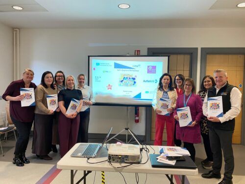 This is a photo of a group of people standing around a projector screen. They are all holding a copy of a new resource called Let's Explore Behaviour Parent Guide. Commissioner Chris Quinn is standing at the end of the row of people on the right-hand side.