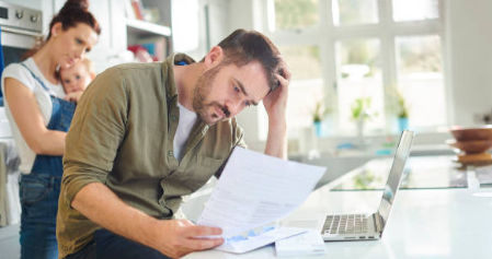 This is a photo of a parent reading a letter at a kitchen table. There is a woman behind him holding a baby looking worried.