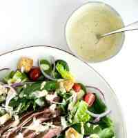 salad on a plate with caesar salad drizzled on top sitting next to a bowl of caesar dressing with a spoon inside.