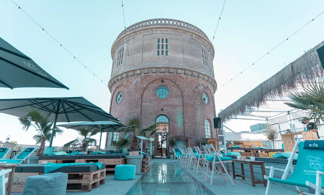 Eine Terrasse mit Liegen und Tischen unter Schirmen vor einem runden Backsteinturm