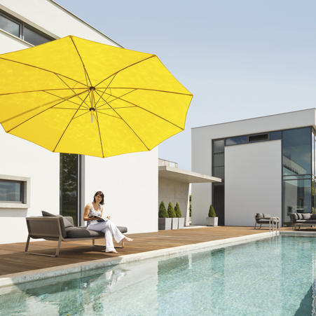 Eine Frau vor einem weißen Gebäude sitzt auf einer Sonnenliege am Pool. Ein gelber MAY DACAPO Sonnenschirm spendet Schatten.