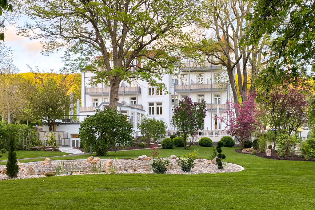 Laudensacks Parkhotel & Retreat mit weißen SCHATTELLO Schirmen Ein Garten mit Bäumen und geschwungenen Wegen neben einer Terrasse mit Sonnenschirm