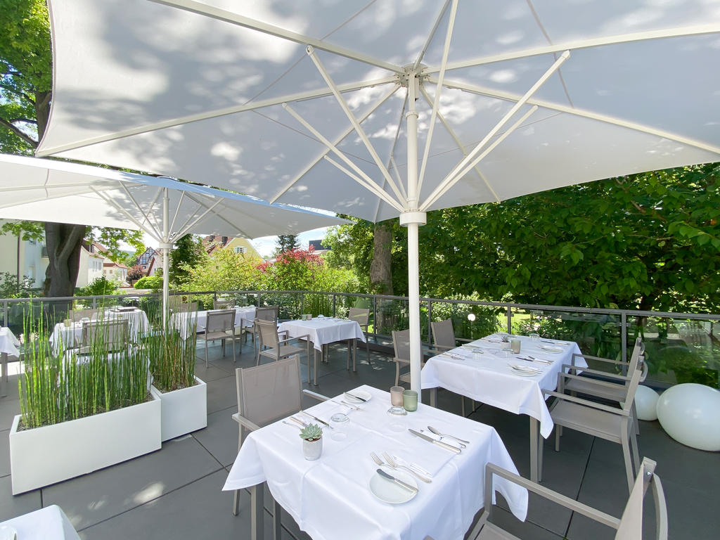 Laudensacks Parkhotel & Retreat mit weißen SCHATTELLO Schirmen Ein Garten mit Bäumen und geschwungenen Wegen neben einer Terrasse mit Sonnenschirm