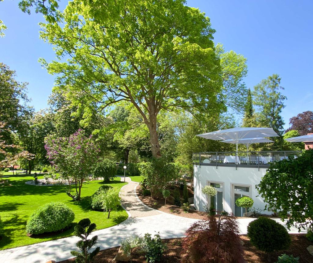 Laudensacks Parkhotel & Retreat mit weißen SCHATTELLO Schirmen Ein Garten mit Bäumen und geschwungenen Wegen neben einer Terrasse mit Sonnenschirm