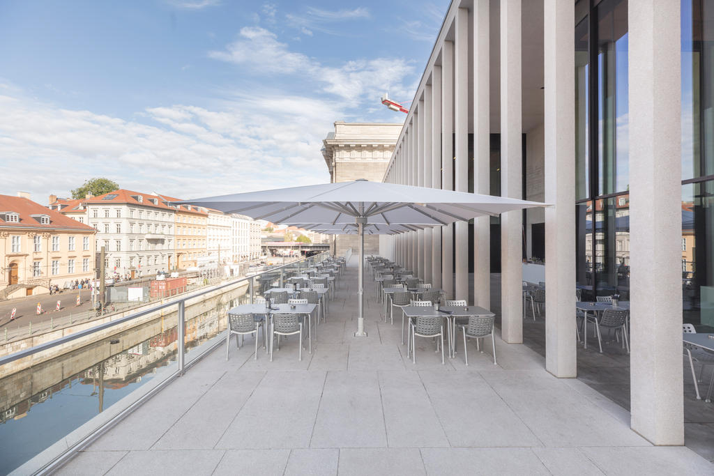 Museumsinsel, James-Simon-Galerie weißen ALBATROS Schirmen Eine moderne Außenterrasse mit weißen Sonnenschirmen, Tischen und Stühlen entlang eines Kanals neben einem Gebäude